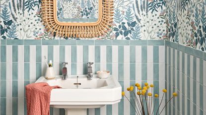 Small bathroom splashback ideas are always useful to know. Here is a bathroom with blue floral wallpaper, green and whote tiles, a white sink, and a rattan mirror