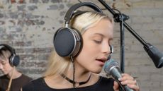 A young white woman with blonde hair is singing into a microphone while wearing Audeze LCD-S20 over-ear headphones.