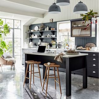 a kitchen with black cabinets along the wall with black panelling halfway up the wall, marble grey floor tiles, a black freestanding kitchen island with white marble top and a full length glass window with black frame letting in light