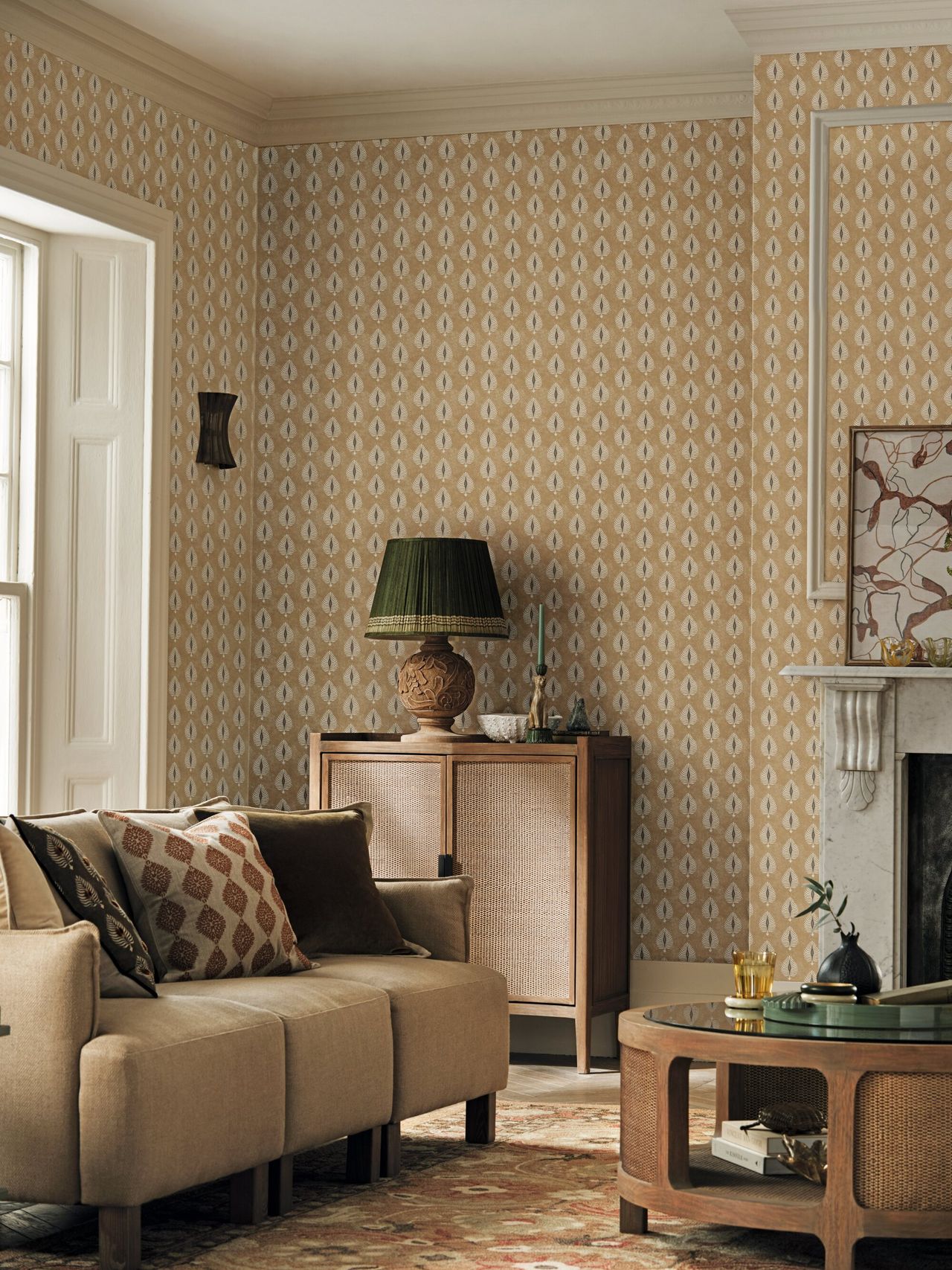 Image of a living room with a warm, beige color palette. There is a patterned wallpaper on the wall, a small beige sofa, a round wooden coffee table, and a wooden console with a green lamp on it.