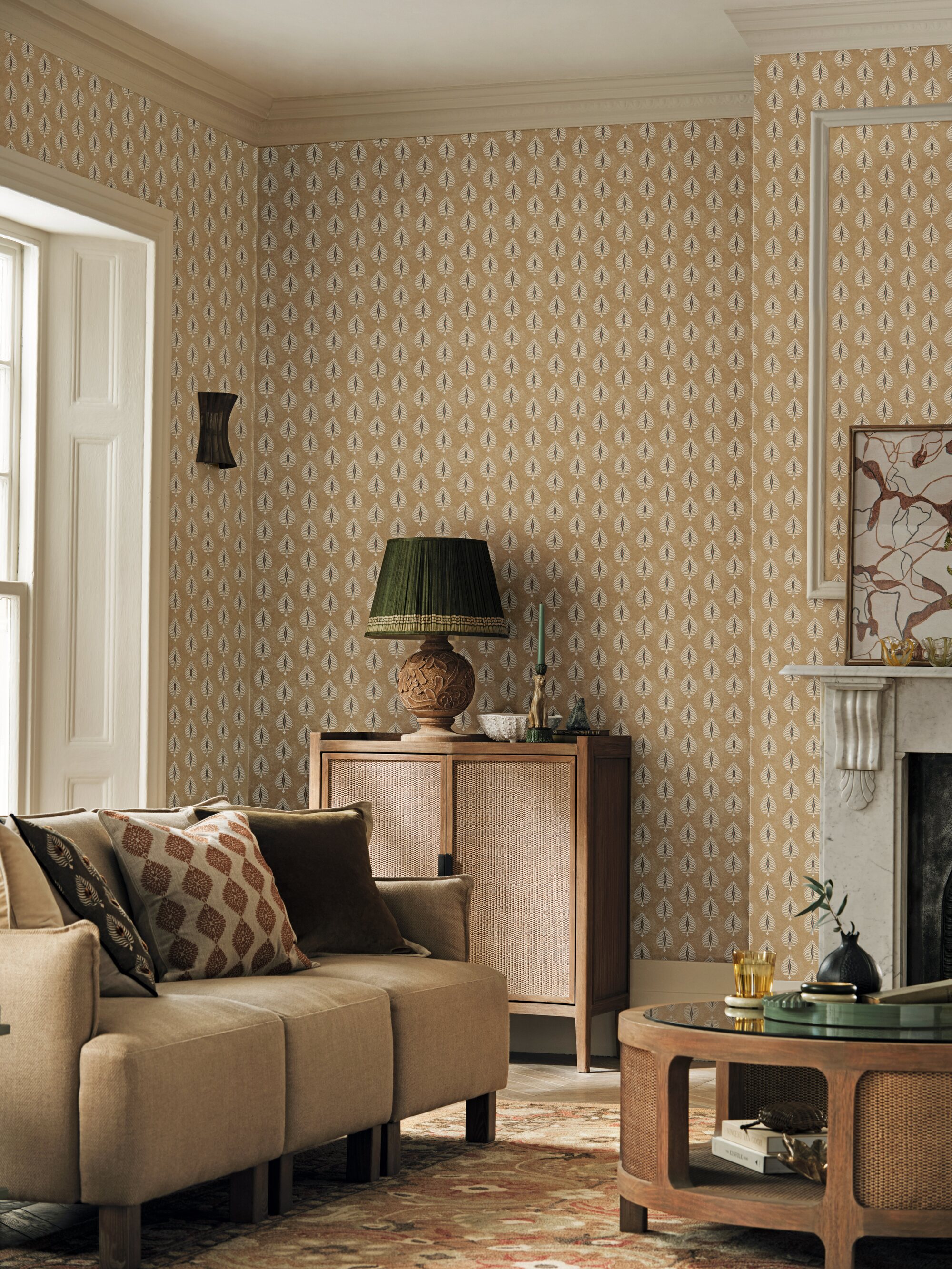 Image of a living room with a warm, beige color palette. There is a patterned wallpaper on the wall, a small beige sofa, a round wooden coffee table, and a wooden console with a green lamp on it.