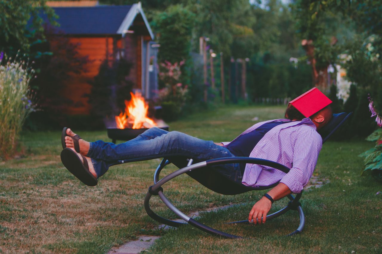 A man in a camp rocking chair sleeps with a book on his face near an outdoor fire.