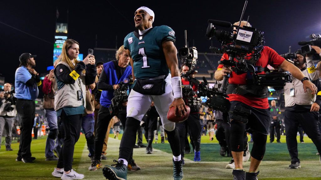 A smiling Jalen Hurts walks off the field surrounded by cameras after a Philadelphia Eagles win.