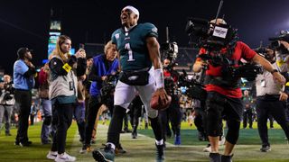 A smiling Jalen Hurts walks off the field surrounded by cameras after a Philadelphia Eagles win.