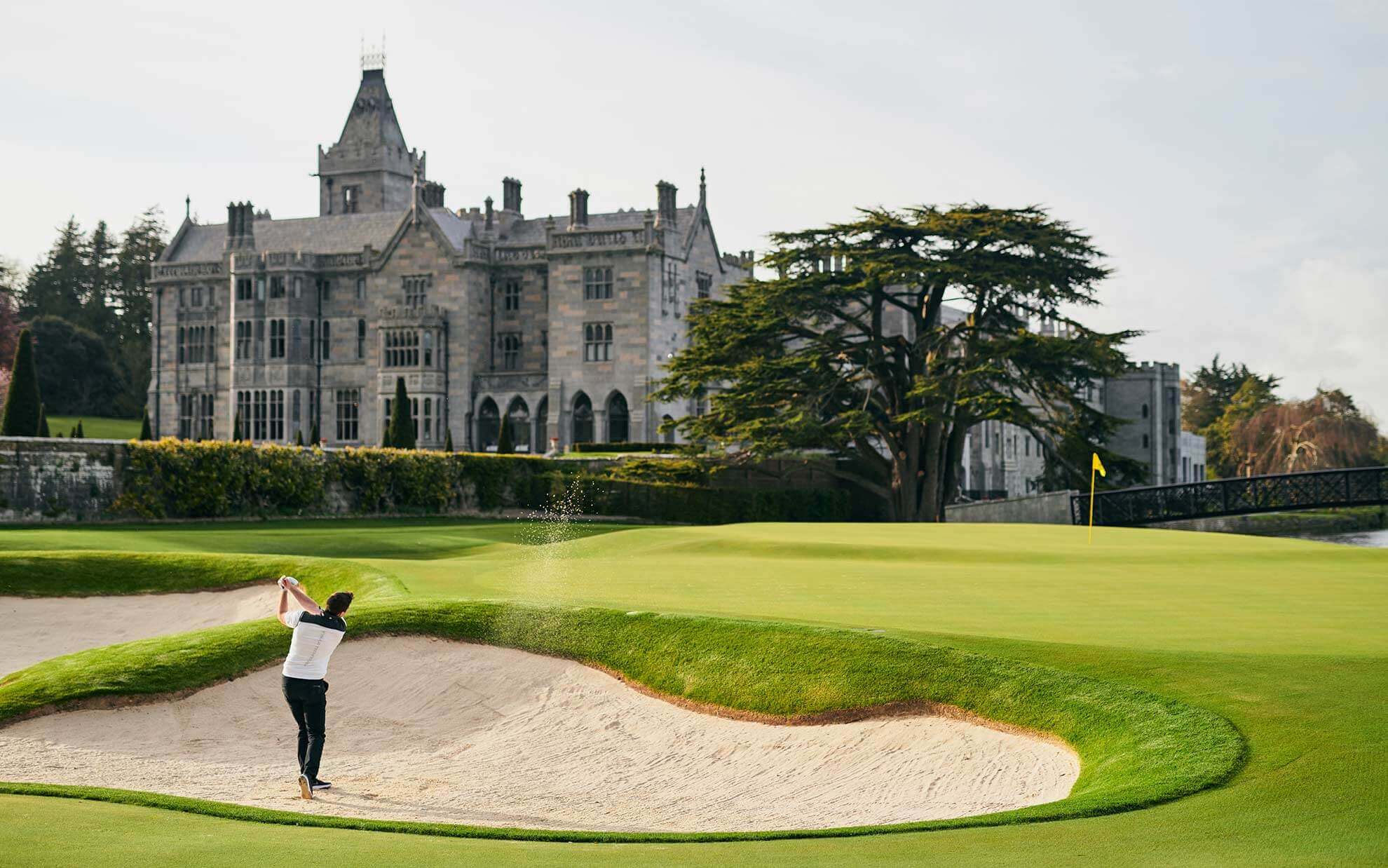 Man hitting a golf ball out of a bunker infront of Adare Manor hotel