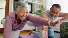 A man and woman practice Pilates at home, smiling. They're on their hands and knees, with one harm outstretched in front and one leg outstretched behind. 
