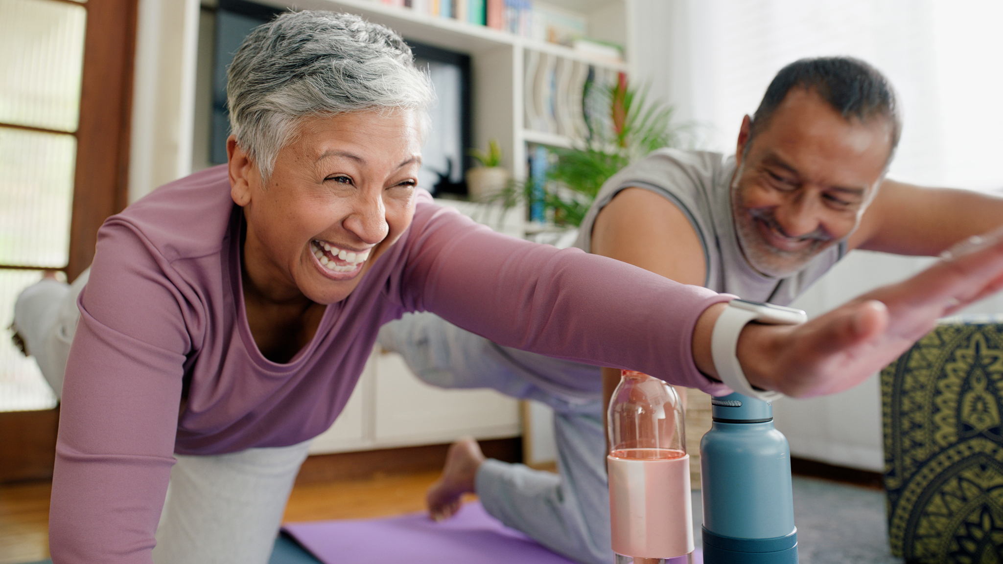 A man and woman practice Pilates at home, smiling. They're on their hands and knees, with one harm outstretched in front and one leg outstretched behind. 