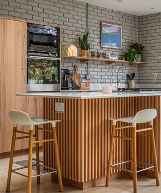 A kitchen island with a white surface with a pink mug and black hob on it, a wooden base, two white and wood bar stools on the sides of it, and a gray brick wall behind it with wooden shelving and cabinet units