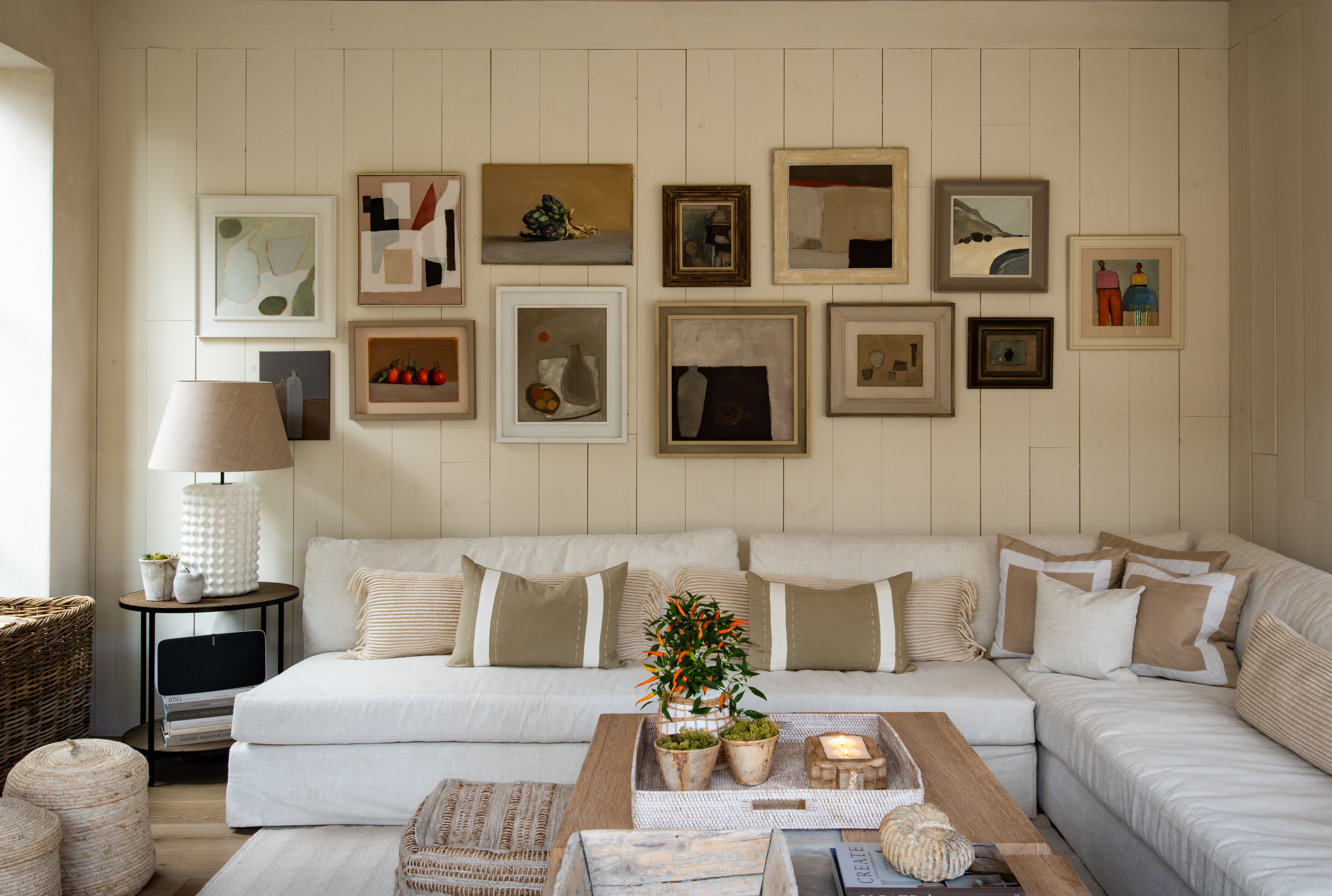 A sitting room with white clad walls covered in paintings of different shapes and sizes, and a white l-shaped sofa 