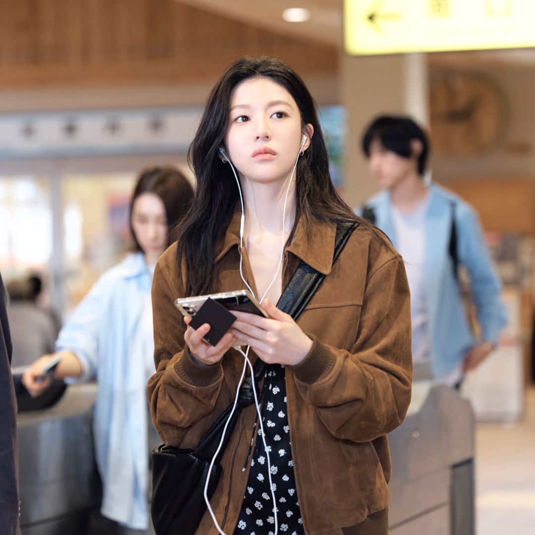 A woman (Go Youn-jung as Cha Mu-hee / Do Ra-mi) wears wired headphones and holds a foldable smartphone as she stands in a lightly crowded train station. A still from the K-drama 'Can This Love Be Translated?'