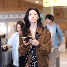 A woman (Go Youn-jung as Cha Mu-hee / Do Ra-mi) wears wired headphones and holds a foldable smartphone as she stands in a lightly crowded train station. A still from the K-drama 'Can This Love Be Translated?'