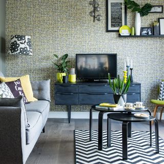 A living room with a micro-print wallpaper, a grey sofa and a TV on top of a media console