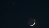 A thin crescent moon shines in a black sky with Venus appearing as a bright star-like object to its upper left.