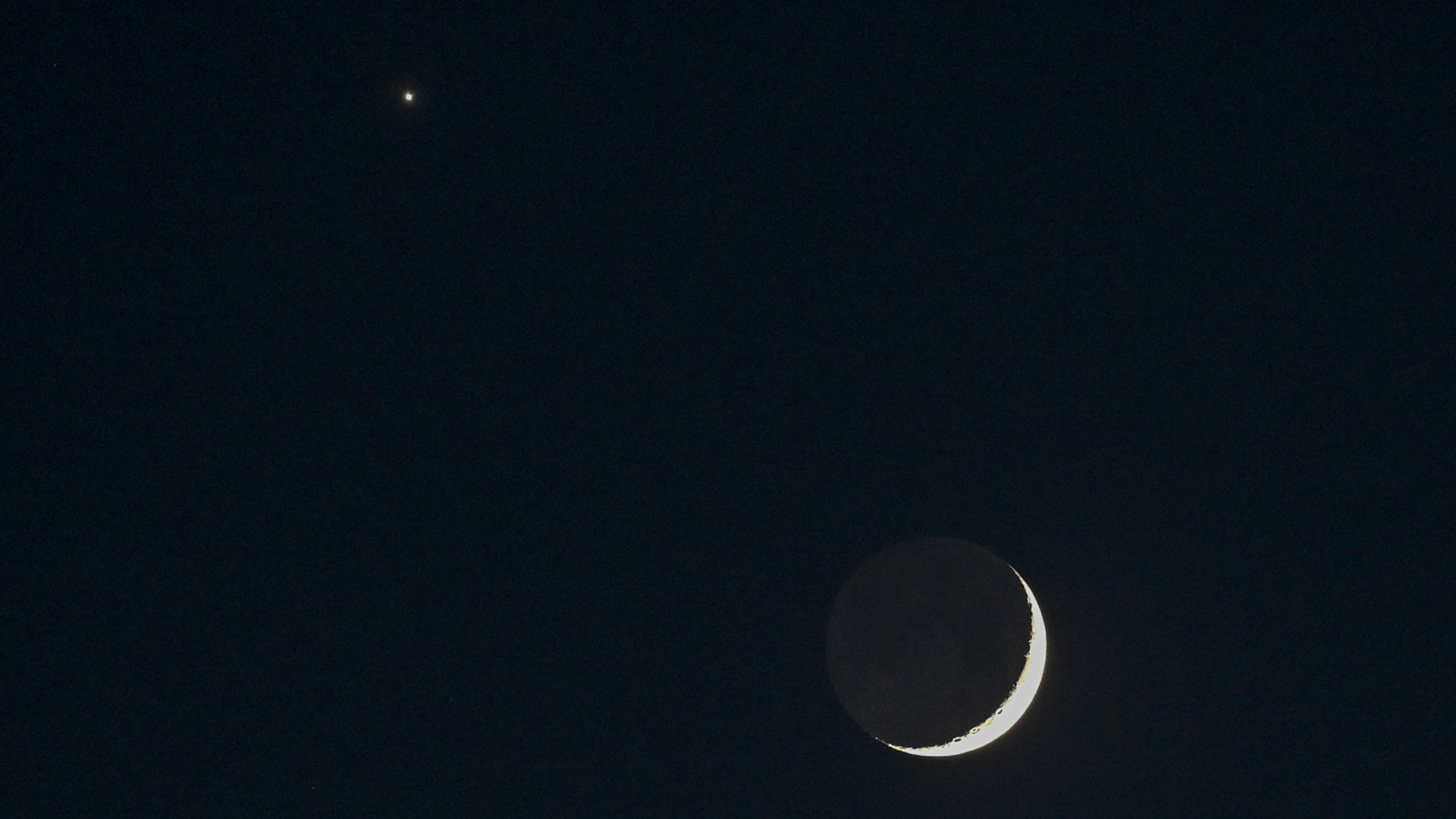 A thin crescent moon shines in a black sky with Venus appearing as a bright star-like object to its upper left.