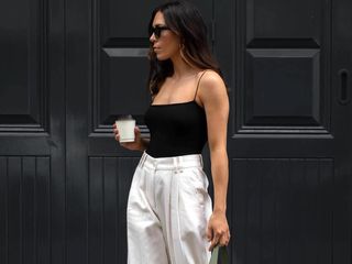 British influencer Jessica Skye poses outside a black paneled building in London wearing black sunglasses, a black tank top with spaghetti straps, and white trousers.