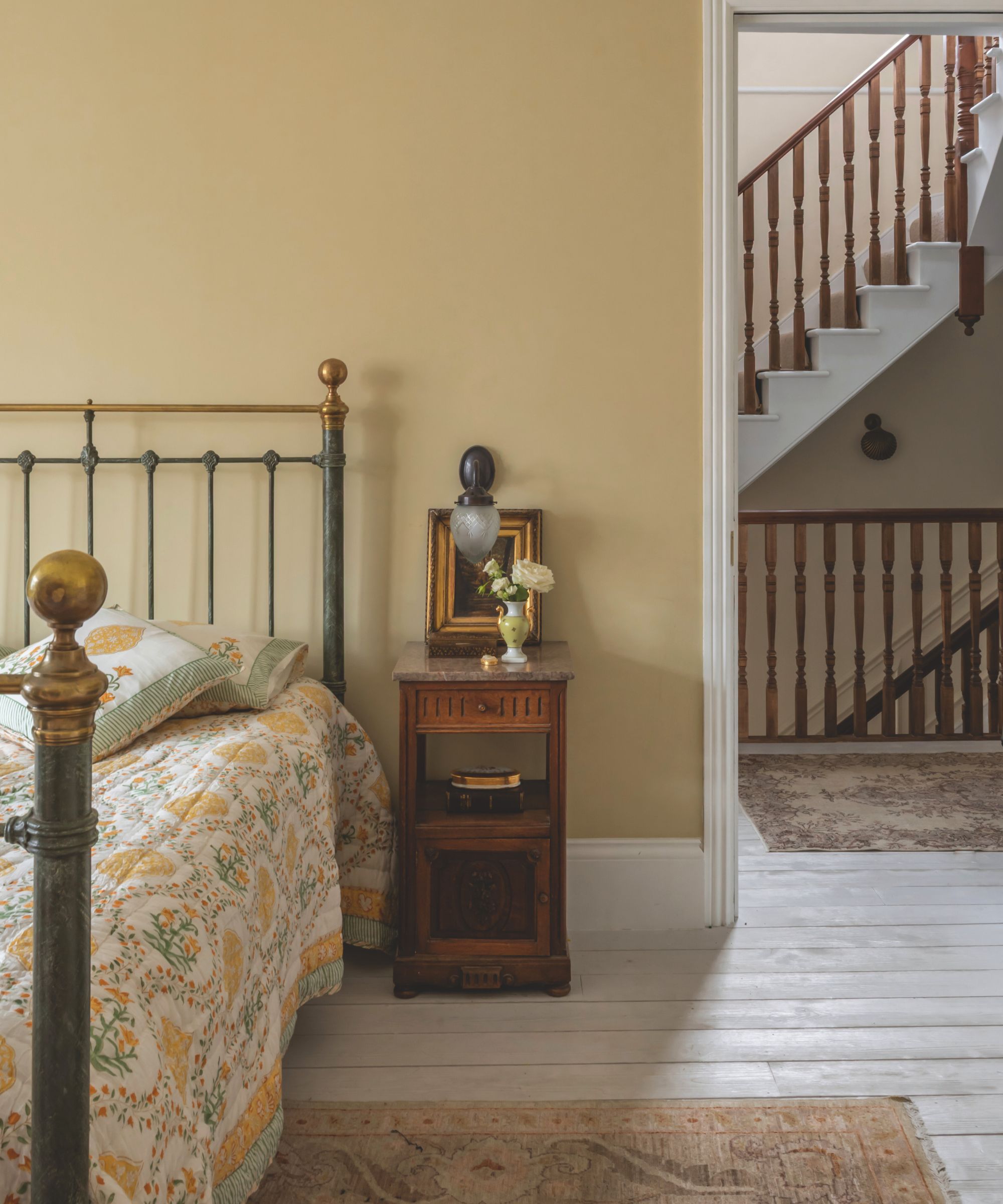 A yellow painted bedroom with a iron bed, and an antique small bedside table. A doorway view of stairs.