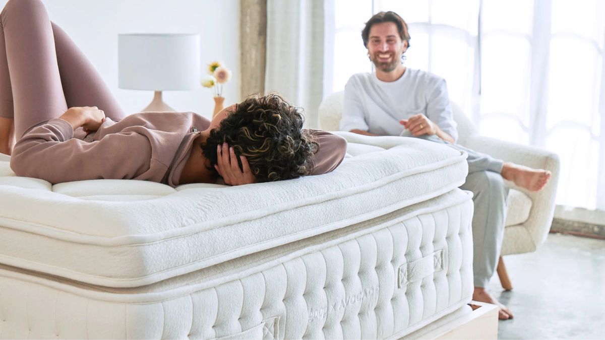 A woman lying on the Goop Avocado mattress with her husband sat on a chair smiling back at her