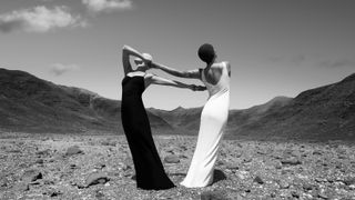 Two women in elegant black and white gowns, holding hands, stand in a rocky landscape, under a clear sky and the crescent moon above