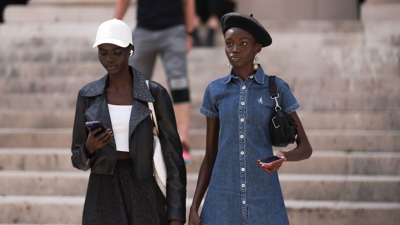 Two women walking down the steps outside a show at Paris fashion week, one is wearing a black skirt, black leather jacket and a white cap, the other is wearing a denim mini dress and a black beret