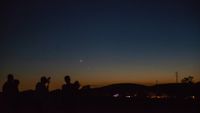 people are silhouetted against a predawn sky as the sun begins to turn the horizon hazy orange and bright stars and planets are still visible above against a deep inky blue sky.