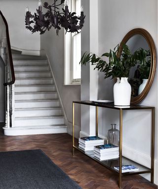 Elegant entryway with dark wood flooring, black rug, metallic tiered console table, decorated with vases, books, ornaments and flowers, white staircase, black leaf chandelier