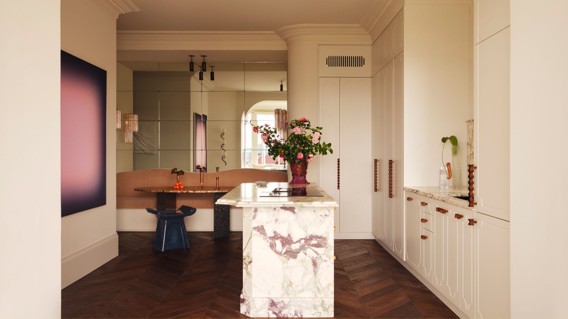 An open kitchen and dining space with a marble island, a vase of flowers, a brown banquette, a curved dining table, and framed purple art