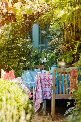 An outdoor dining area with a colorful patterned tablecloth, wooden chairs with peach and blue throw pillows, and a pergola with green vines above it