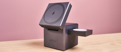 The Anker 3-in-1 Cube with MagSafe sits on a light-colored wooden surface with a light pink background. The charging pad has been raised, revealing some of the AirPods charging area at the back. The Apple Watch charging pop-out it sticking out to the right.