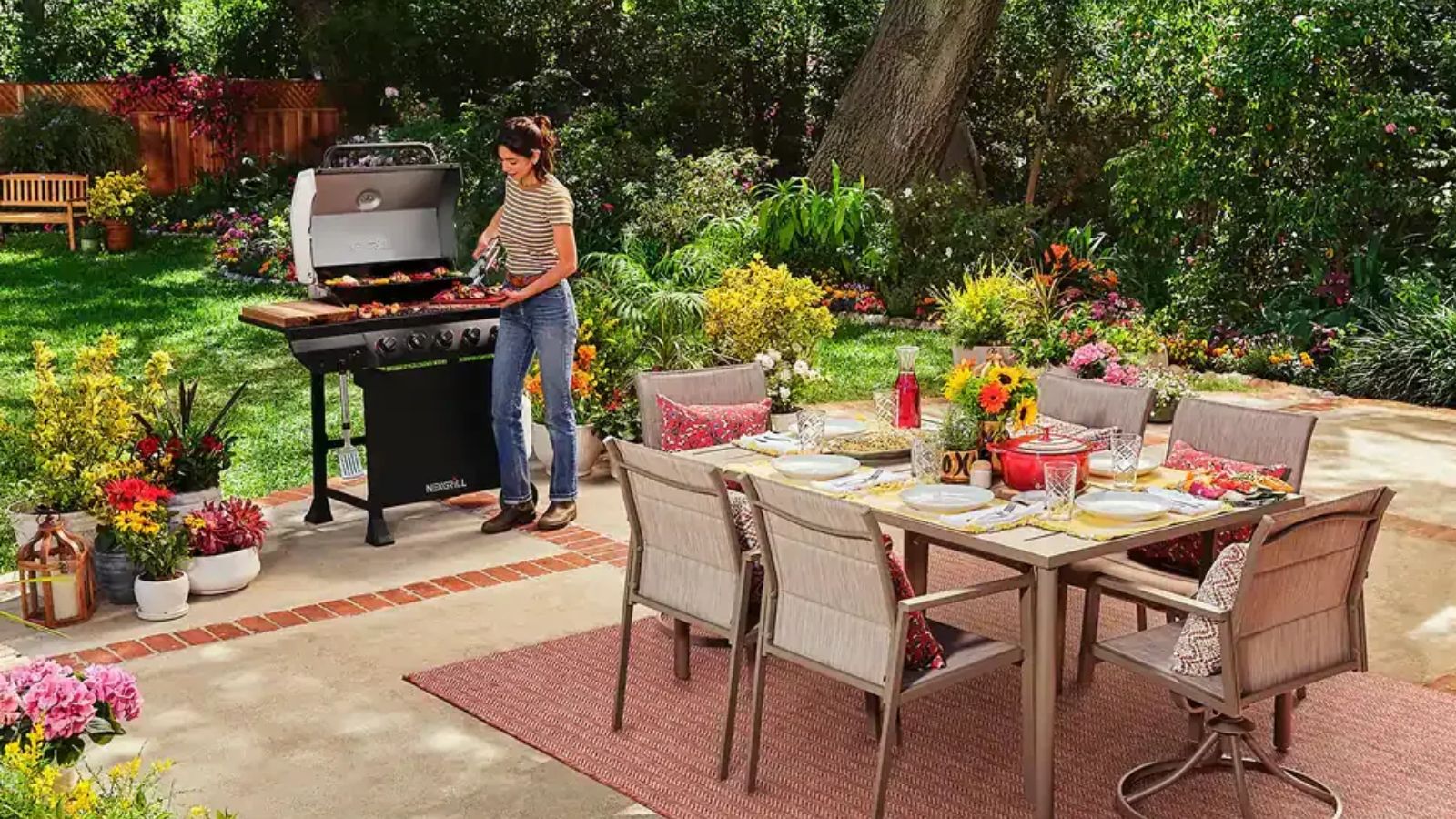 Large patio with outdoor rug and dining table and chairs, woman standing over a BBQ to te left, greenery and flowers in the background