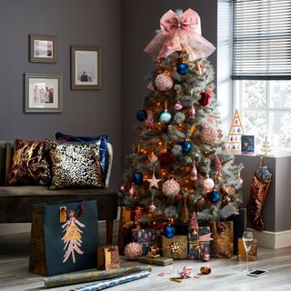 A Christmas tree covered in colourful decorations and a large pink bow on the top situated in a grey living room.