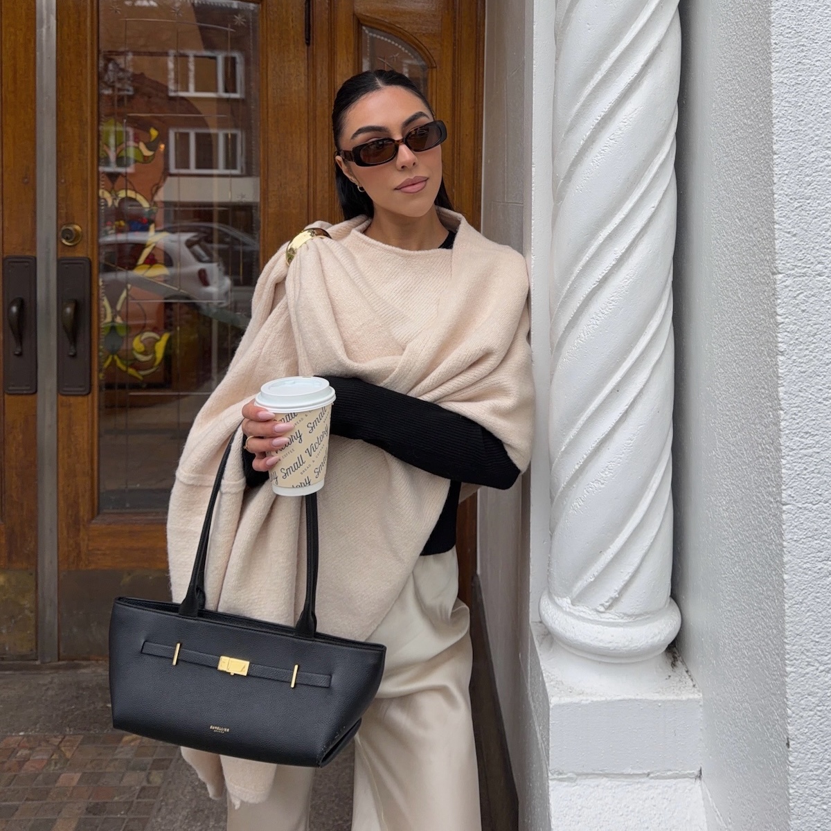 A well-dressed woman holding a bag and a coffee while leaning against a building