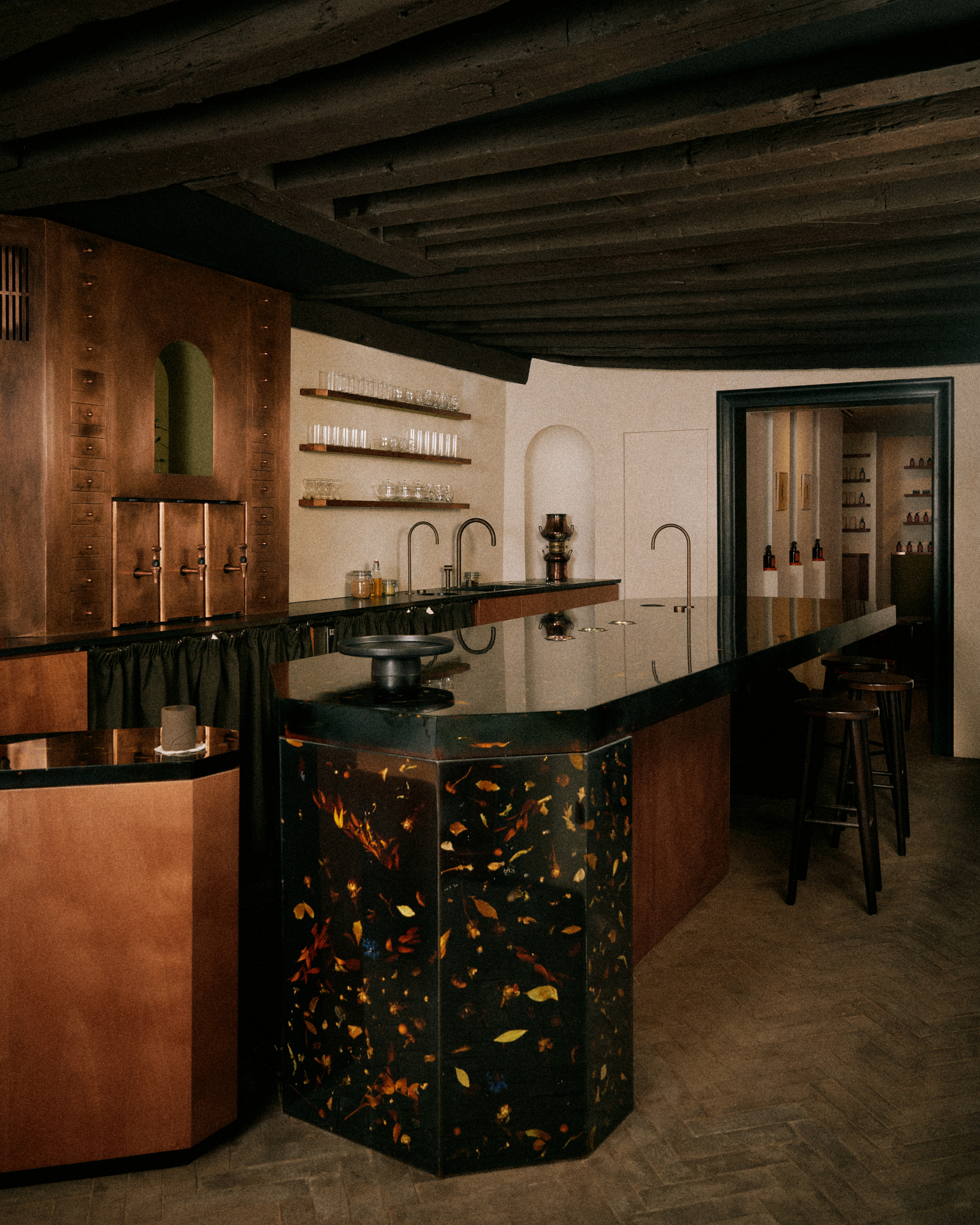 A naturally lit teahouse bar and dining room cast from resin filled with florals, decorated with bronze surfaces mixed with wood and beige plasters, along with wooden stools.