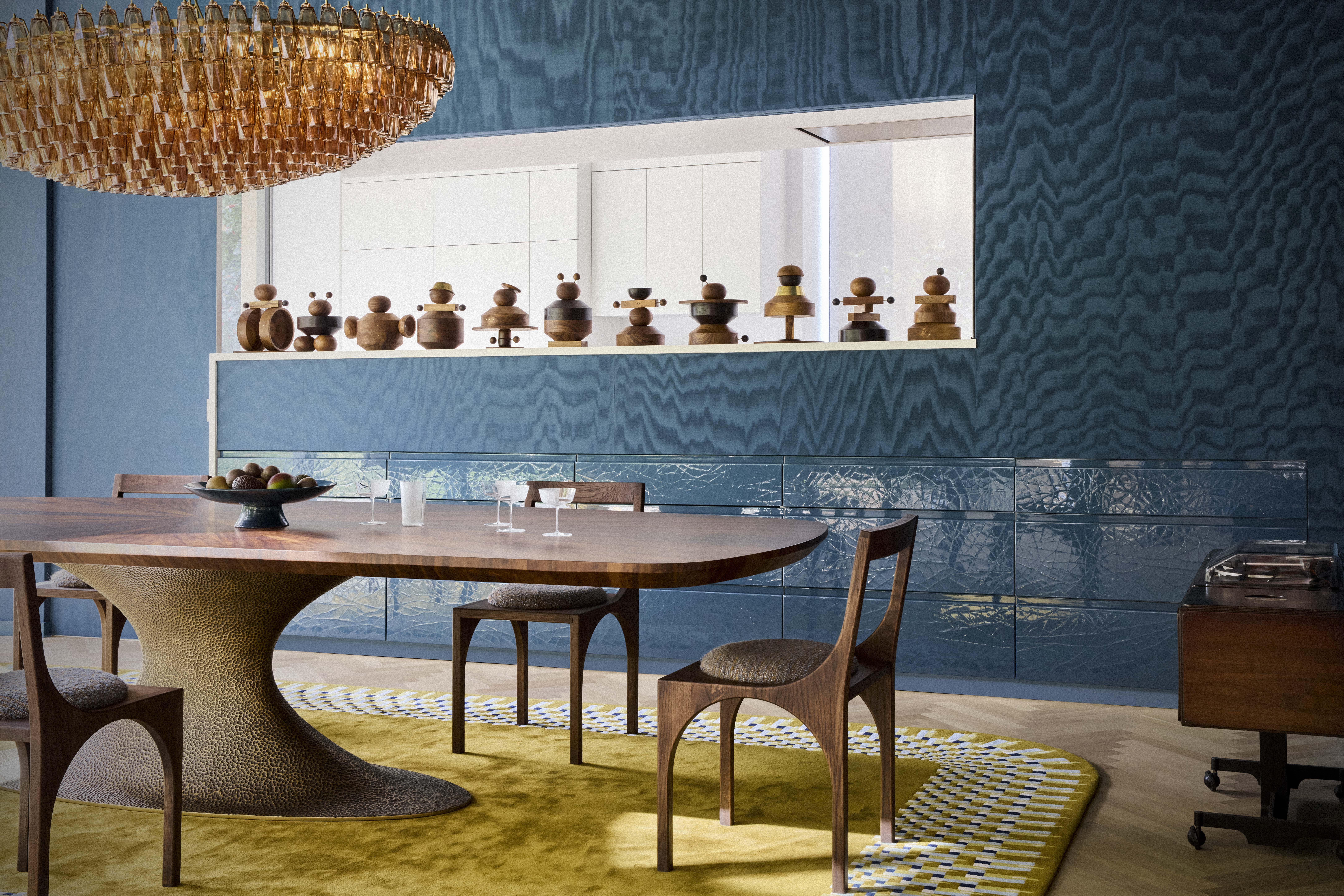 A large dining room with a blue wallpaper, a sculptural dining table unit, a chandelier and a yellow carpet