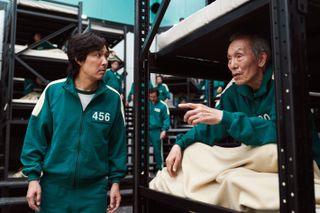 A man (Lee Jung-jae) listens to another man (Oh Young-soo) who sits in a bunk bed with a blanket over his lap, in 'Squid Game.'