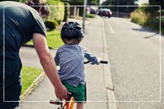 Father holding on to bike seat to steady the bike, while his helmet-wearing child learns to ride