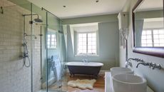 A bright bathroom with a glass shower screen, black roll top bath with sheepskin rug below a bright window, two matching circular porcelain basins with silver faucets and a wooden framed mirror