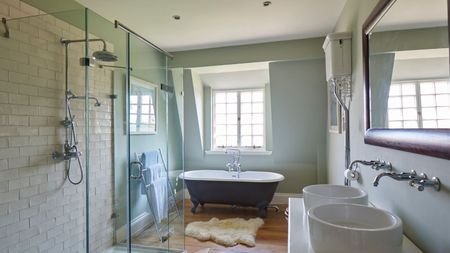 A bright bathroom with a glass shower screen, black roll top bath with sheepskin rug below a bright window, two matching circular porcelain basins with silver faucets and a wooden framed mirror