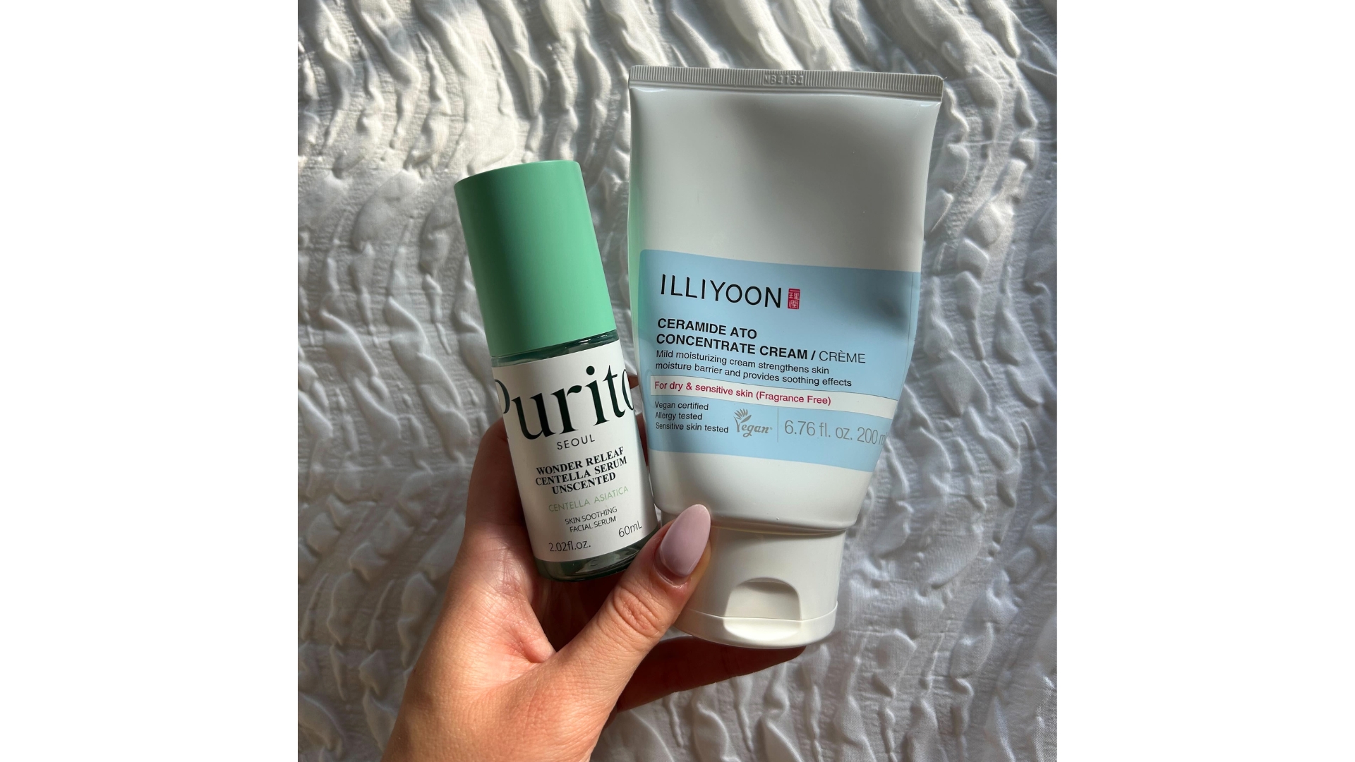 Hand holding Illiyoon Ceramide Ato Concentrate Cream and Purito Centalla Serum against white textured bedsheets