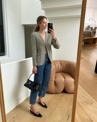 female fashion influencer Anouk Yve poses for a mirror selfie with a low curly ponytail wearing a neutral strong shoulder blazer, straight-leg jeans, black shoulder bag, and black slingback flats