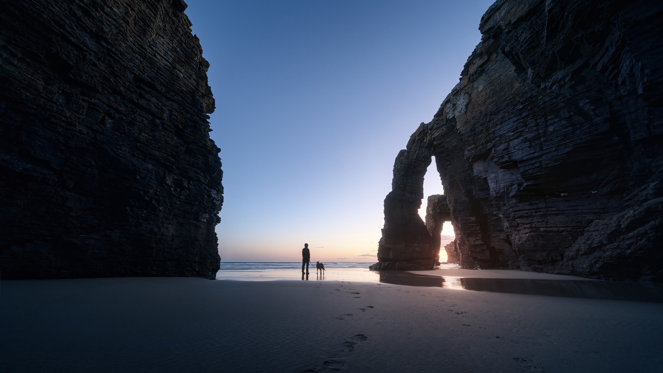 stunning sunset show with vast rocky structures either side of a man and his dog looking out to the setting sun over the sea.