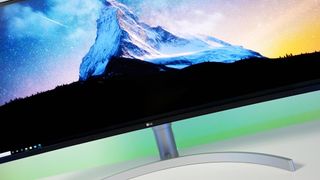 Close-up view of an LG ultrawide curved computer monitor mounted on a silver curved stand. The screen displays a vivid desktop wallpaper featuring a sharp, snow-covered mountain peak set against a starry sky transitioning from blue to yellow. A Windows taskbar is visible along the bottom left corner.