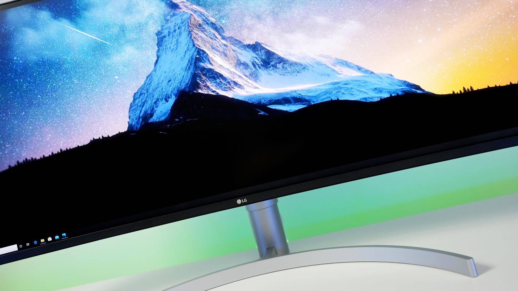 Close-up view of an LG ultrawide curved computer monitor mounted on a silver curved stand. The screen displays a vivid desktop wallpaper featuring a sharp, snow-covered mountain peak set against a starry sky transitioning from blue to yellow. A Windows taskbar is visible along the bottom left corner.