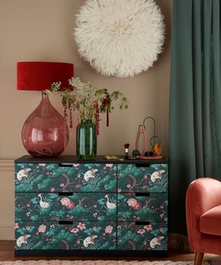Set of drawers covered in patterned wallpaper with red table lamp on top