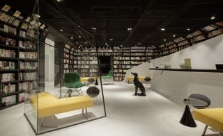 Interior view of Wheat Youth Arts Hotel, Hangzhou, China featuring white floors, wall-to-wall bookshelves filled with books, green and yellow seating, round tables, spotlights and a black dog sculpture chained to the black and white reception desk