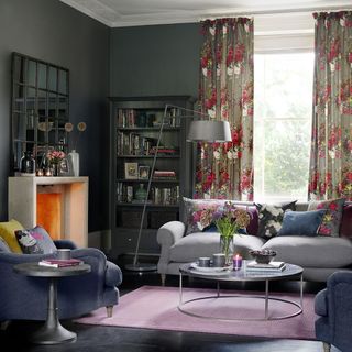 grey living room with floral curtains next to a wooden book shelf with a grey sofa with floral pillows and a circle shaped coffee table on top of a purple rug