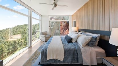 Bedroom with floor to ceiling windows overlooking mountainside trees, with ceiling fan, hardwood floors, wood paneling on the wall and layered bedding.