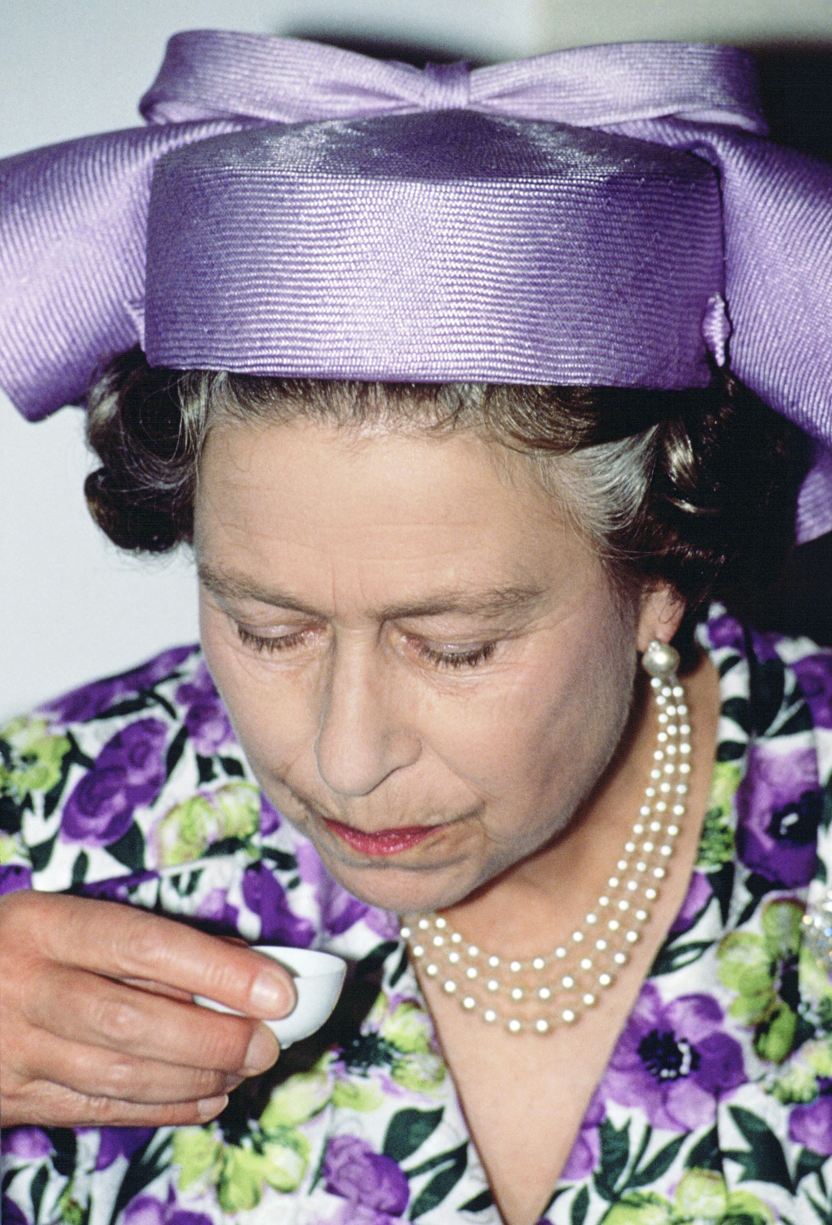 Queen Elizabeth wearing a purple floral dress and sipping tea