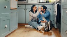 A young couple smile and high-five while sitting together on the floor of their kitchen.