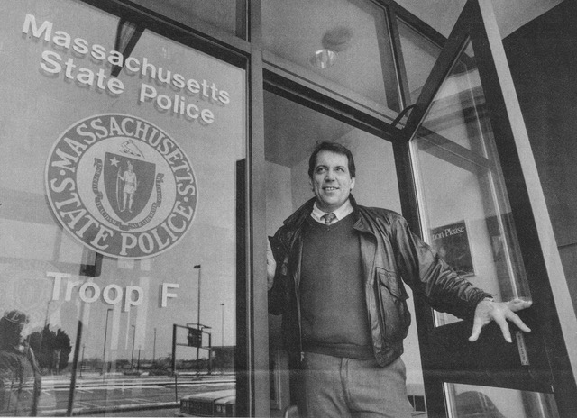 A male Massachusetts state trooper stands in a doorway and poses for a photo