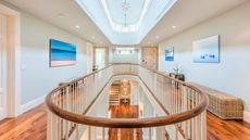 Grand hallway with coastal accents such as wicker benches and seaside art on walls. The stairs are in view in the background, with an oval hallway railing and gallery view over the lower floor. The floors are hardwood and the ceiling is brigh with a matching oval window gallery